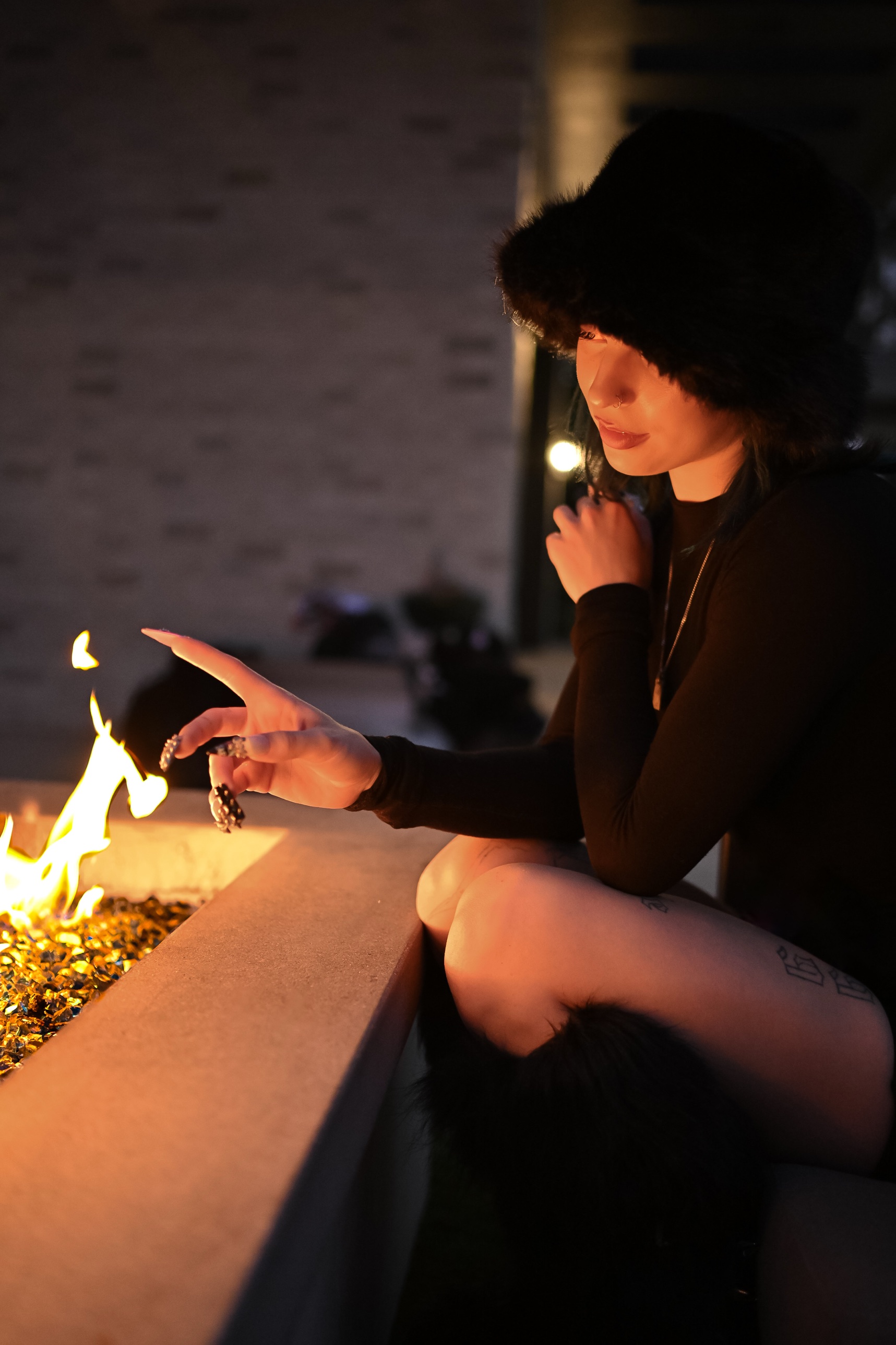 Woman in a dark fur hat and black outfit reaching toward the flames of an outdoor fire pit at night