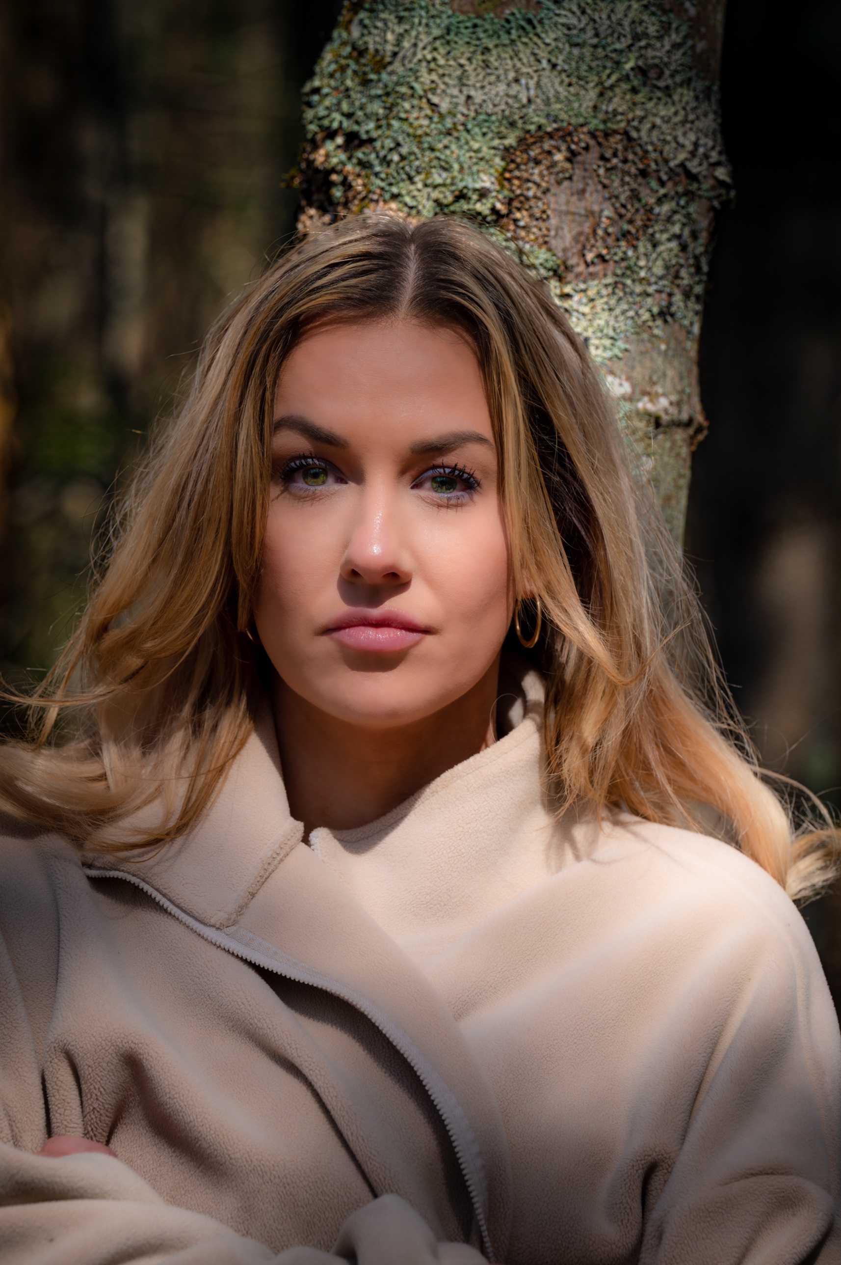 Close-up portrait of a woman in a tan coat standing against a lichen-covered tree trunk