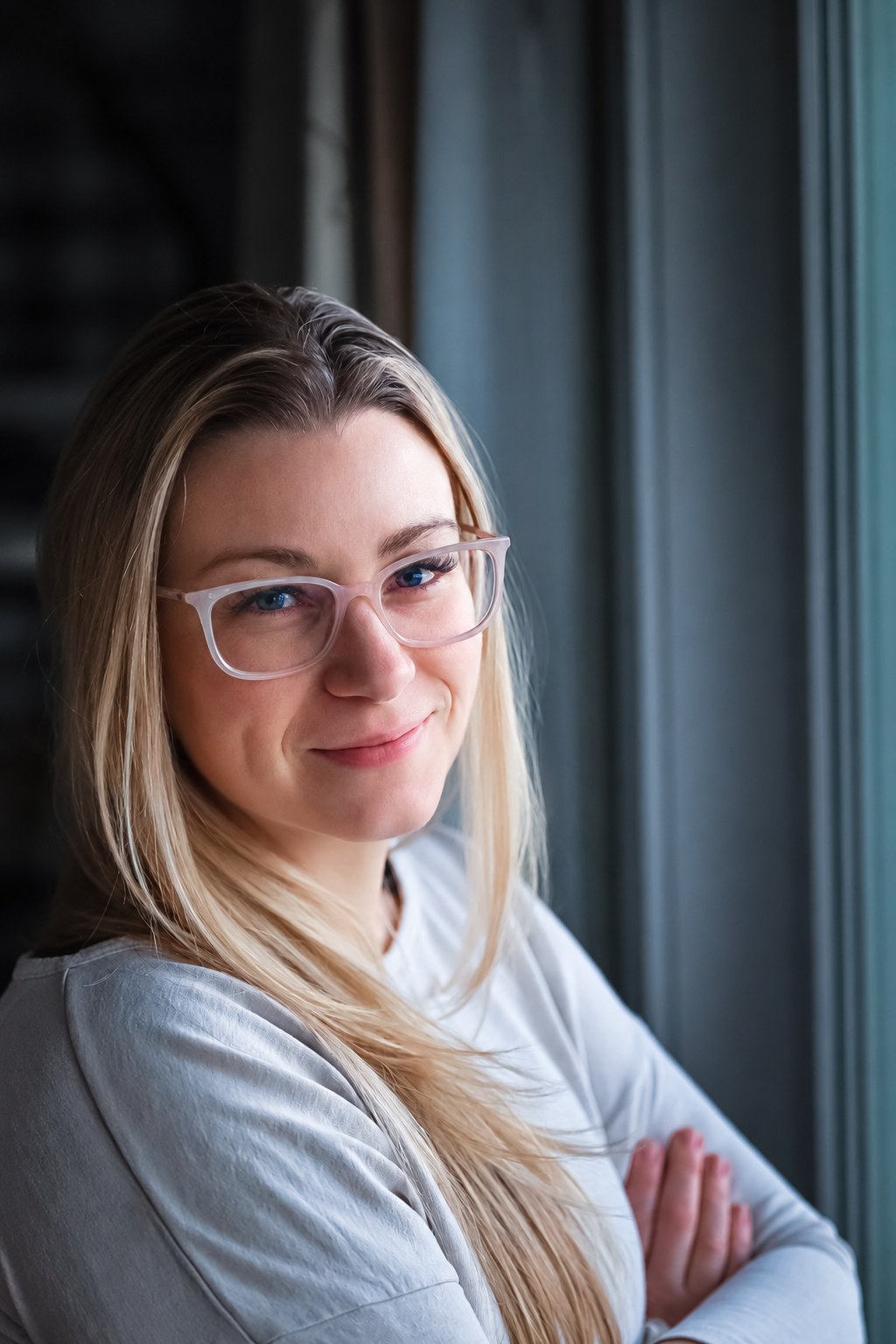 Woman with glasses smiling softly by a window, arms crossed