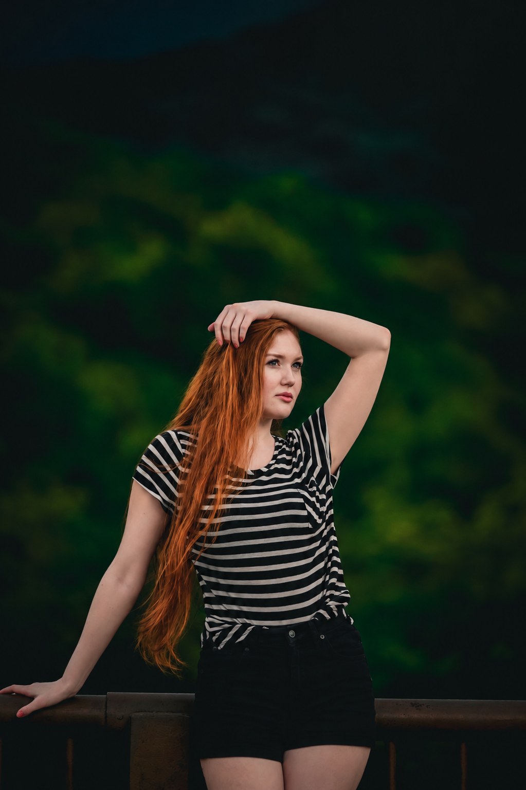 Redheaded woman in a striped shirt leaning against a railing against a dark green background