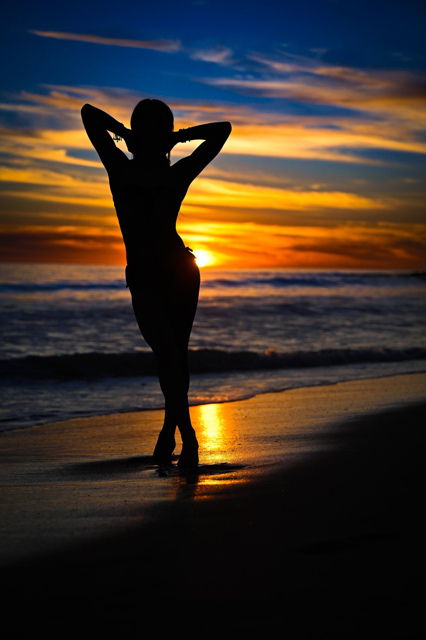 Silhouette of a woman from behind, hands in her hair, against a beach sunset