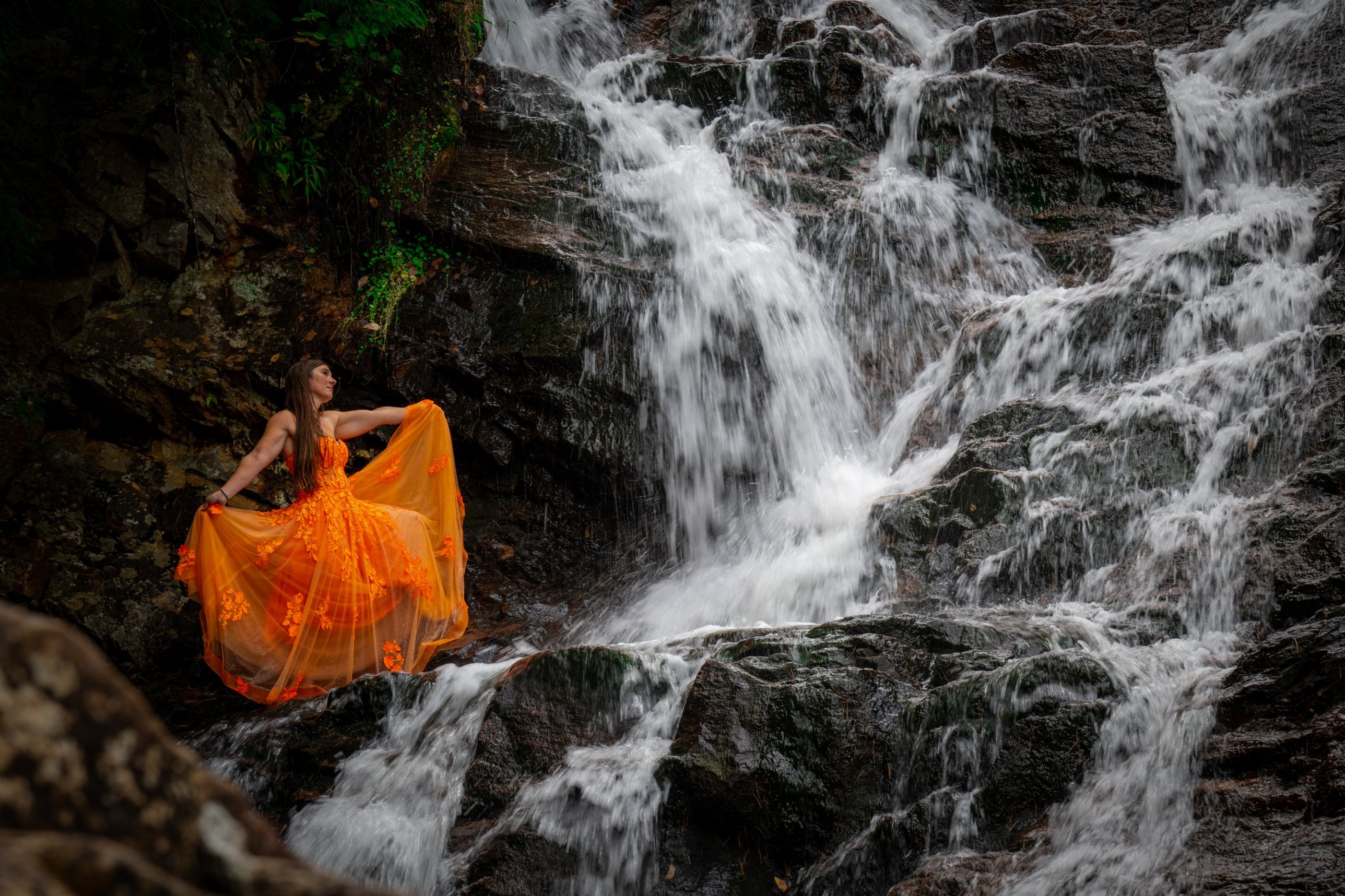 Woman in a flowing orange gown standing on rocks beside a cascading waterfall
