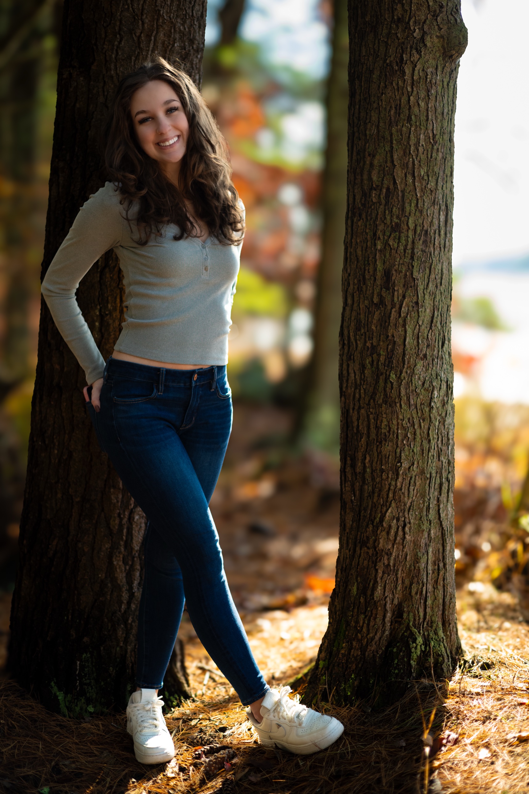 Senior portrait of a young woman smiling and leaning between two tree trunks in a fall forest