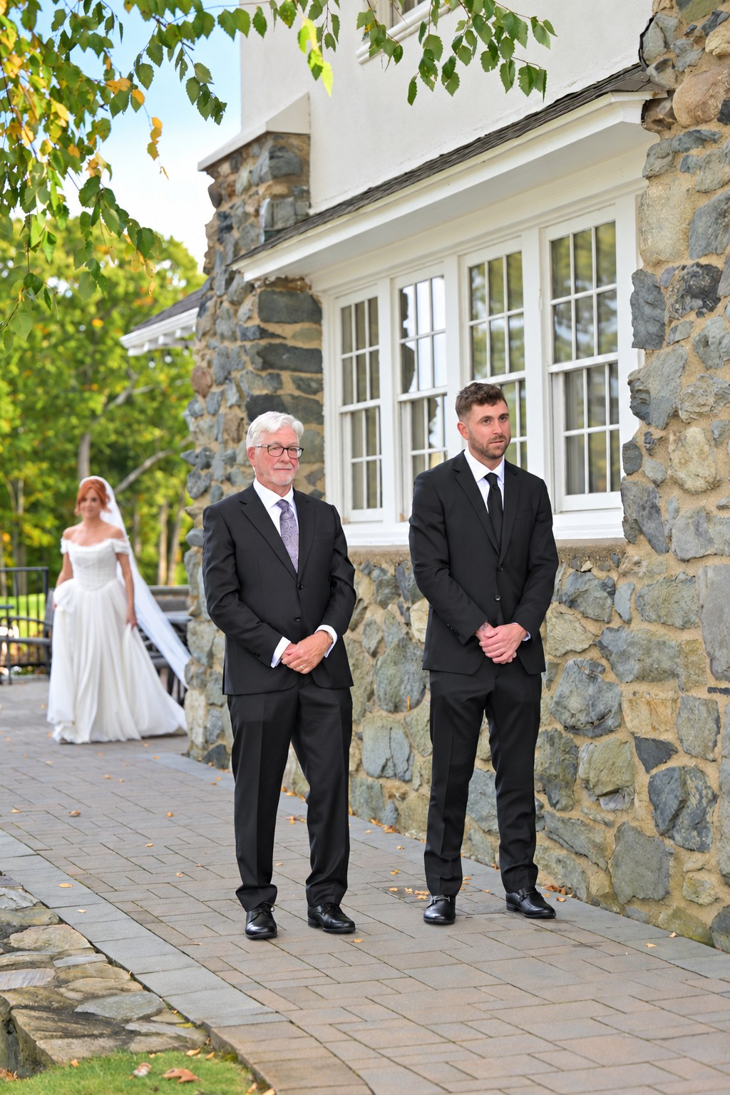 Groom and father of the bride waiting outside a stone building as the bride approaches in the distance
