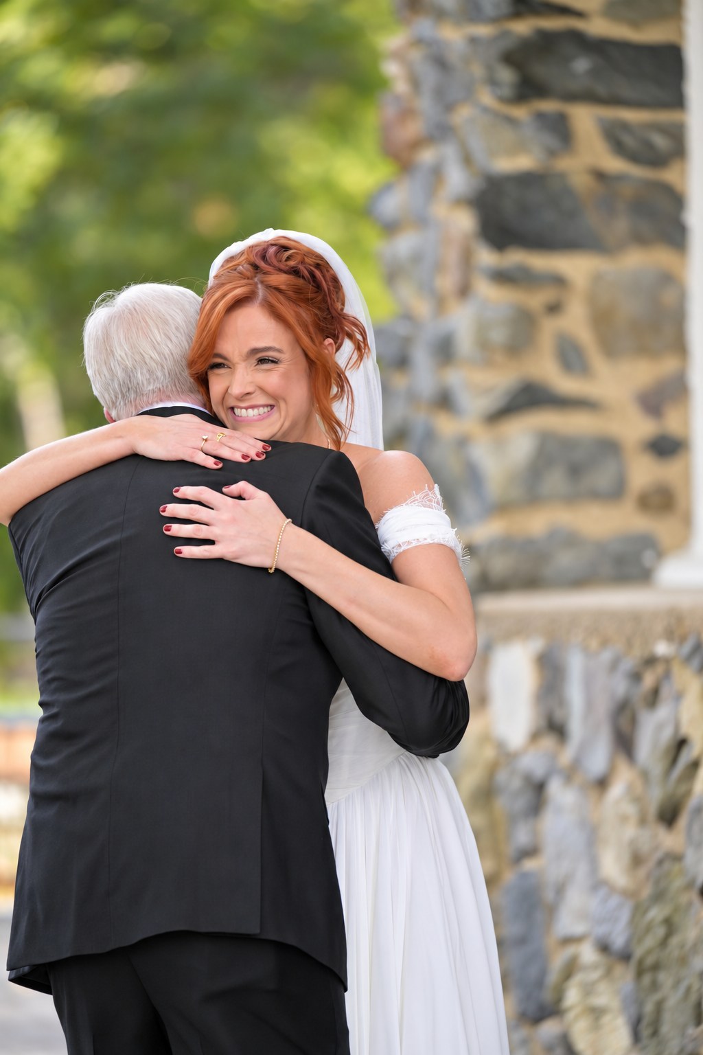 Bride laughing and hugging her father beside a stone wall