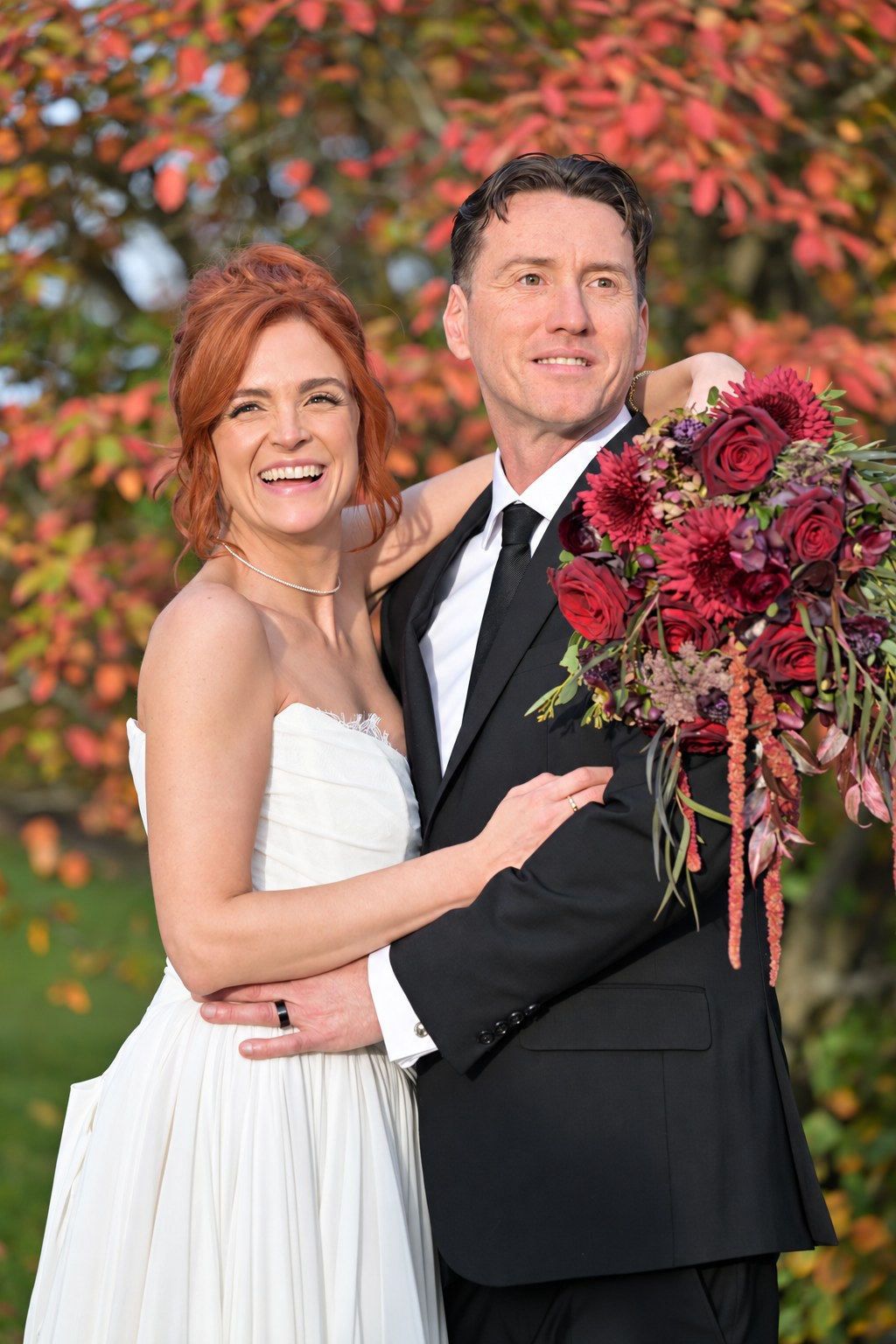 Bride and groom smiling together in front of fall foliage, holding a deep red bouquet