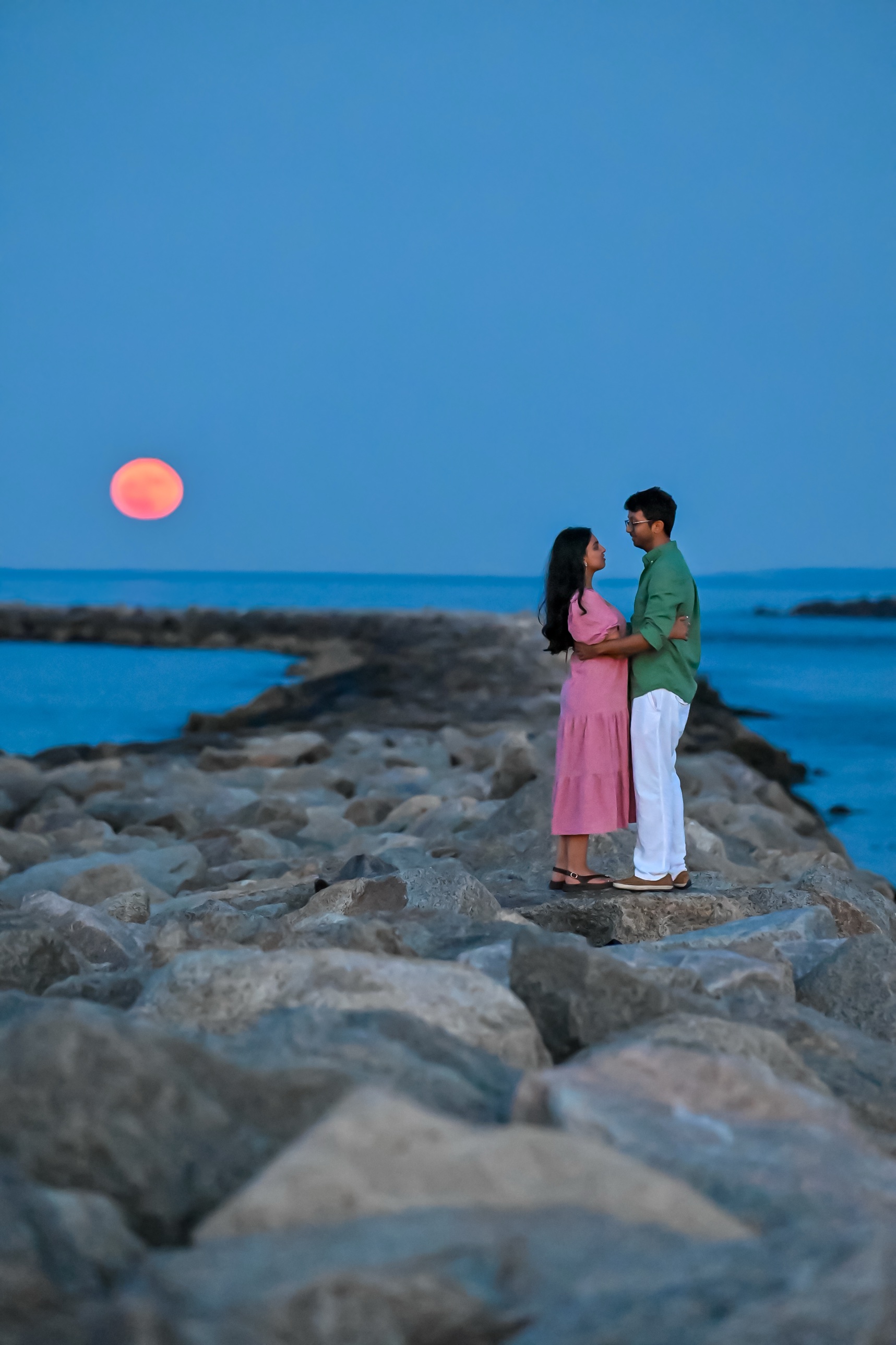 Engaged couple embracing on a rocky jetty as a full moon rises over the ocean